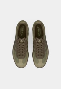 Olive green sneakers with a mix of mesh and suede materials, featuring brown laces and three signature stripes on the sides. Flat rubber sole.