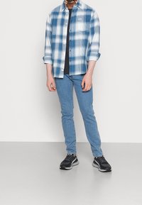 Chemise en flanelle à carreaux bleus et blancs, avec une poche classique, associée à un t-shirt sombre et un jean bleu clair. Chaussures de sport noires avec semelles grises.