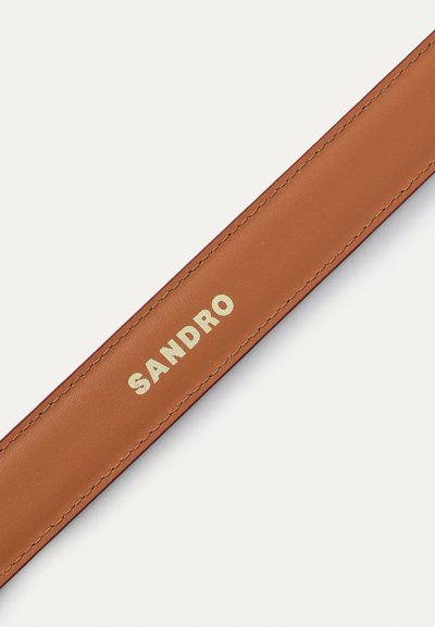 Kerge pruuni nahast rihm kuldse embossitud "SANDRO" logoga, sile tekstuur, minimaalne õmblus ja ristkülikukujuline disain.