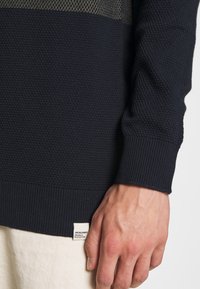 Maglione lavorato a maglia blu navy con un motivo testurizzato, polsini e orlo a coste. Presenta un'etichetta piccola con il nome del marchio vicino al fondo.