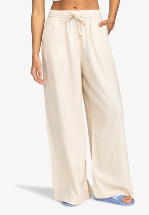 Pantalon large crème à taille coulissée porté par une personne debout, assorti de sandales bleu ouvert à l'avant.