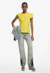 Gelbes Oberteil mit strukturierten Blumenakzenten, hellblaue Jeans mit dunklen Patches, hält eine blaue Jeansjacke. Plateausandalen runden den Look ab.