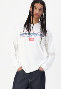 Hvit langermet skjorte med "new balance" logo i blått og rødt, sammen med mørke denimjeans. Enkel snitt og avslappet passform.