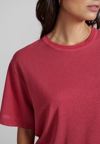 Červené tričko s krátkým rukávem vyrobené z měkké, texturované látky s kulatým výstřihem. Vyznačuje se uvolněným střihem a minimálními švy.