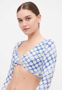Long-sleeve crop top in blue with yellow floral pattern, featuring a deep V-neck and gathered center detail. Smooth, stretchy fabric.