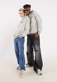 Zwei Personen tragen übergroße graue Hoodies mit einem kleinen Grafik auf der Brust, kombiniert mit blauen Jeans und zerrissenen schwarzen Jeans.