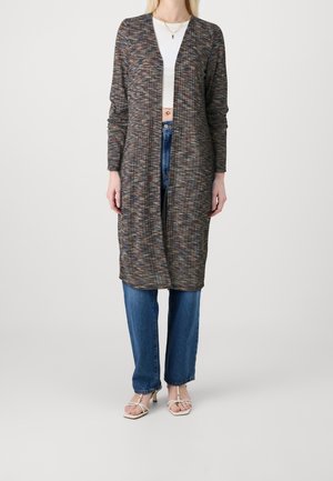 Vrouw die een meerkleurige lange cardigan draagt over een wit cropped topje, blauwe spijkerbroek en witte sandalen met hakken, staand tegen een effen achtergrond.
