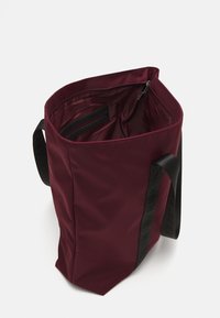 Sac fourre-tout bordeaux en nylon avec une poche zippée noire à l'intérieur et de larges sangles noires. Forme structurée avec une grande ouverture.