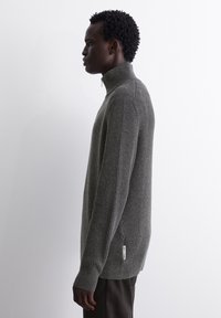 Grau gerippter Strickpullover mit hohem Kragen, langen Ärmeln und einem seitlichen Etikett. Der Stoff wirkt strukturiert und gemütlich.