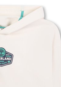 Felpa bianca con un accento teal, caratterizzata da una toppa rotonda con un albero e il testo "Timberland", maniche corte e una texture morbida.