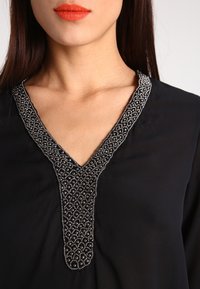 Blusa nera con scollo a V decorato da intricati dettagli in perline argento e un motivo testurizzato lungo il colletto.