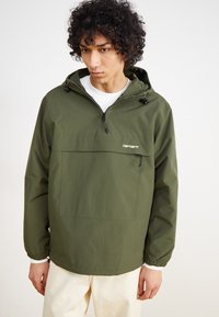 Olivgrüner Windbreaker mit Kapuze, Frontreißverschluss und elastischen Bündchen. Verfügt über eine große Fronttasche und ein Logo auf der Brust.