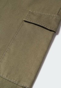 Gros plan sur une manche en tissu vert olive avec une poche rectangulaire cousue et un détail de sangle en tissu assorti.