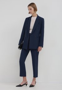 Completo blu navy con giacca doppiopetto, pantaloni aderenti e scarpe col tacco a punta nera. Top chiaro testurizzato sotto.