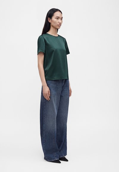 Femme aux longs cheveux foncés, vue de profil, portant un haut en satin vert foncé à manches courtes, un jean bleu à coupe large et des chaussures noires pointues.