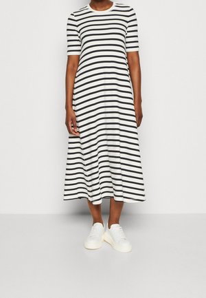 Personne portant une robe midi à rayures horizontales noires et blanches avec des manches courtes et des baskets blanches, debout devant un fond uni.