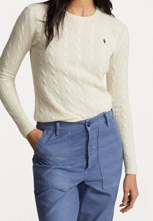 Kobieta ubrana w kremowy sweter w warkocze z logo Ralph Lauren i wysokim stanem niebieskie spodnie, jedna ręka w kieszeni, na jednolitym tle.