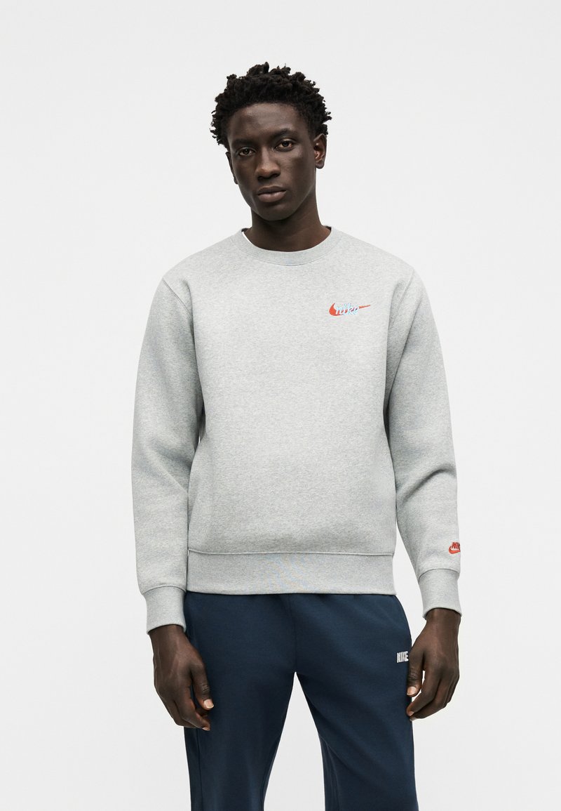 Young man with short curly hair wearing a light gray crewneck sweatshirt and navy blue pants with small red and white Nike logos.