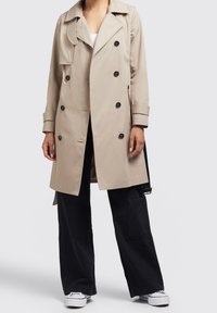 Beige trenchcoat med en dubbelknäppt design, svarta knappar, breda slag och belagd midja. I kombination med svarta vida byxor och vita sneakers.