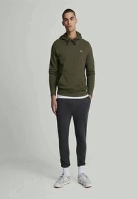Sweatshirt verde com capuz e bolso canguru, combinado com calças de moletão cinza escuro e ajustadas. Realçado com um pequeno logo bordado.