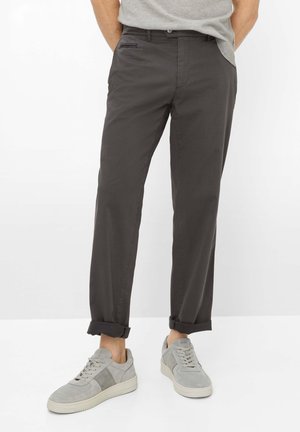 Mann trägt hochgekrempelte dunkelgraue Hose und hellgraue Sneakers, steht vor einem schlichten weißen Hintergrund.