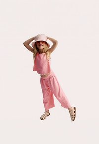 Ensemble deux pièces en tissu rose pâle ; haut court avec ourlet festonné et pantalon ample. Associé à un bob assorti et des sandales.