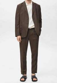 Brązowy garnitur na miarę z usztywnioną marynarką i spodniami o prostych nogawkach, zestawiony z białą koszulką polo i czarnymi sandałami z odkrytymi palcami.