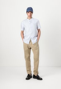 Camicia a maniche corte con bottoni, a righe blu e bianche, pantaloni kaki e scarpe scure, abbinata a un berretto blu con logo.