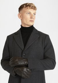 Gants en cuir marron avec un détail de couture portés par une personne dans un manteau en laine gris charbon et un col roulé noir.