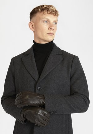 Guanti in pelle marrone con dettagli cuciti indossati da una persona con un cappotto di lana grigio scuro e un dolcevita nero.