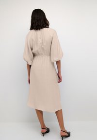 Beiges knielanges Kleid mit Puffärmeln, elastischem Taillenbund, Ausschnitt auf der Rückseite, aus einem strukturierten Stoff gefertigt. Schwarze Absatzsandalen runden den Look ab.