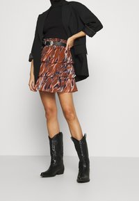 Mini falda marrón con estampado y volantes, combinada con un suéter de cuello alto negro ajustado y una chaqueta negra oversize. Las botas de vaquero negras complementan el atuendo.