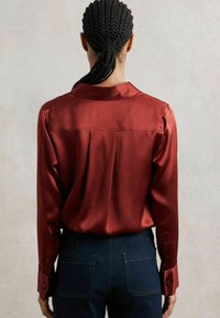 Cămașă din satin roșu cu guler și mâneci lungi, având un pliu în spate și cu manșete butonate, purtată cu blugi de culoare albastru închis.