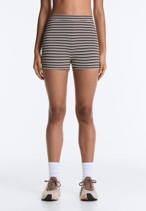 Nő barna-fehér vízszintes csíkos rövidnadrágban, fehér boka zokniban és bézs tornacipőben, karjait lazán oldalt tartva áll.