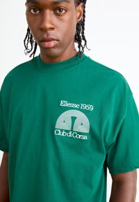 Grøn bomulds t-shirt med rund hals og korte ærmer. Forsiden har hvid broderet logo med buede linjer og tekst.