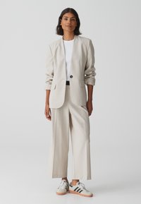 Beige blazer met een enkele zwarte knoop, gecombineerd met beige wijde broek en een wit t-shirt. Schoenen: grijze sneakers met zwarte strepen.