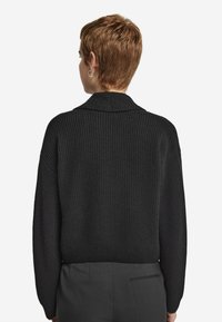 Svart ribbad stickad tröja med krage och långa ärmar. Löst sittande med en kortad nederdel, som visar en texturerad tyg.