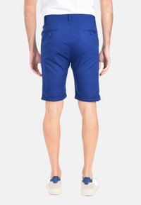 Pantaloni corti blu con una texture liscia, orli arrotolati e doppie tasche posteriori. Indossati con scarpe di colore chiaro.