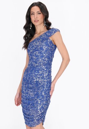 Hosszú sötét hajú nő, aki testhez simuló, egyik vállánál egyvállas, ezüstös fémes csillogású kék ruhát visel, egyik kezét a csípőjén tartva, semleges arckifejezéssel pózol.