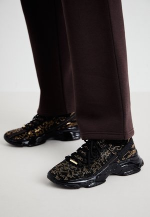 Schwarz-goldene Sneakers mit Muster und dicken schwarzen Sohlen, getragen zu dunkelbraunen Hosen auf einem hellen Boden.