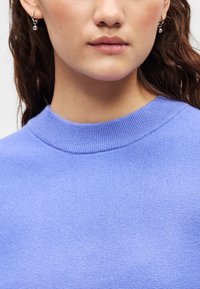 Ανοιχτό μωβ πουλόβερ με ριμπ ύφανση στο στρογγυλό λαιμό και λεία υφή. Κρεμαστά σκουλαρίκια σε χρώμα επιδερμίδας εμφανή, προσθέτοντας λεπτομέρεια στο πάνω μέρος της εικόνας.