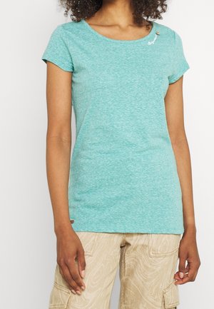 Mujer con una camiseta ajustada de manga corta en color verde azulado claro y pantalones cargo beige con estampado, de pie frente a un fondo blanco liso.