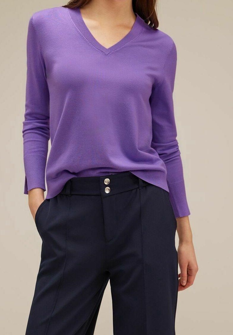 Femme portant un pull violet à manches longues avec col en V, légèrement rentré dans un pantalon taille haute bleu marine à deux boutons, les mains dans les poches.