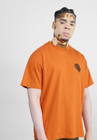 Camiseta de algodón naranja con un corte relajado, que presenta un pequeño logotipo de corazón negro en el área del pecho izquierdo. Diseño simple, mangas cortas.