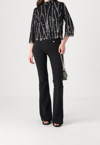 Sort, pailletteret cropped top med trekvartærmer, parret med højtaljede, tætsiddende sorte flared bukser. Har guldfarvede knapper.
