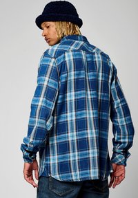 Camisa de franela a cuadros en tonos azules claros y oscuros, con puños abotonados y dobladillo recto. Textura suave, cuello estándar y detalles en el yugo posterior.