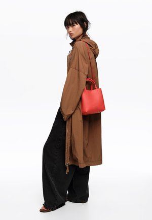 Mujer con una chaqueta marrón grande con capucha, pantalones negros anchos y sandalias, llevando un pequeño bolso rojo colgado del hombro.