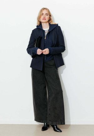 Femme portant un blazer bleu marine à fines rayures sur un pull zippé bleu marine, une chemise rayée, un jean noir large, des bottines noires, tenant une pochette noire.