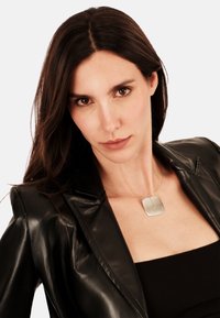 Stříbrný náhrdelník s pendansem na tenkém řetízku, se čtvercovým designem a centrální štěrbinou, nošený přes černý top a koženou bundu.