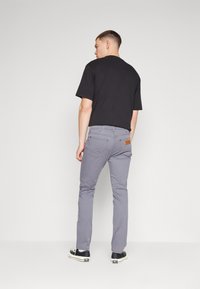 Grijze herenbroek met een slim fit, gemaakt van katoen, vijf zakken, bruine leren label op de achterkant, gecombineerd met een zwart t-shirt en sneakers.